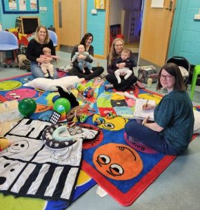 A group of parents sitting on a floor with their babies in their laps. Our Engagement Officer is also with them. The floor is covered in brightly coloured play mats