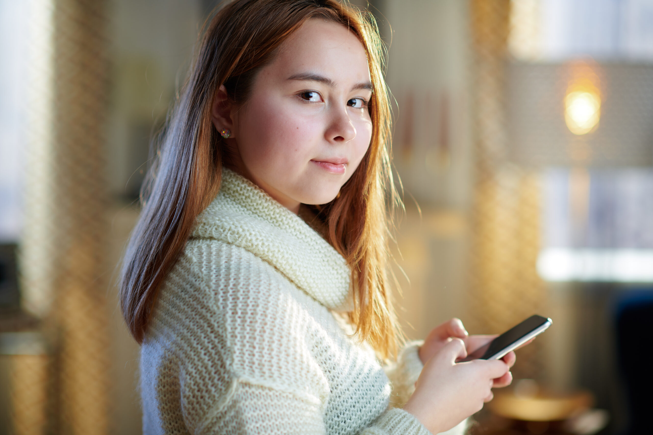 modern teenager girl with red hair in white sweater with smartphone hanging in social media in the modern house in sunny winter day.