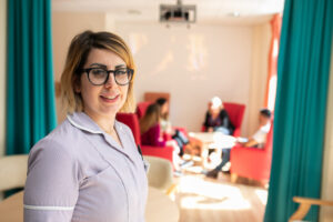 A care home worker is standing at the front of the photo, to the left of centre. In the background are people sat chatting to each other in high backed chairs.