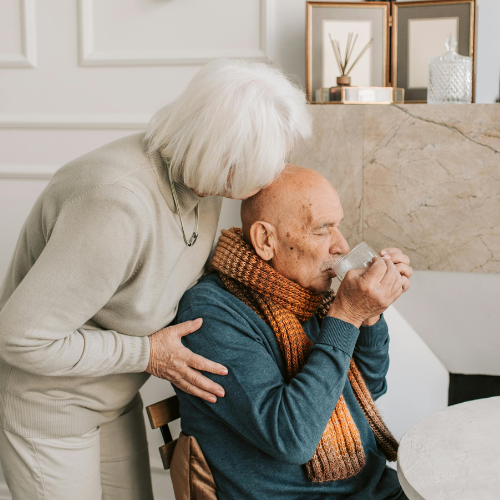 A person is sitting down sipping a drink they are holding with both hands. Behind them a person is toching their arm and leaning into to them.