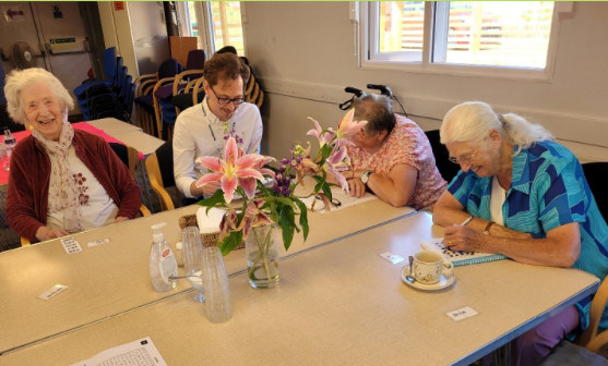 4 people are sat at a table, 3 of them are looking down at papers on the table, one is looking at the camera. All of them are smiling.